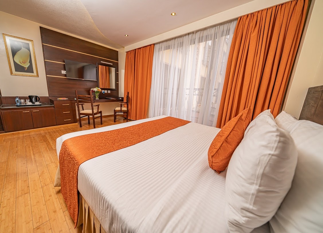 A cozy sleeping area features a spacious bed adorned with an orange throw and decorative pillows. Natural light fills the room through large glass doors, complemented by flowing sheer curtains. A wooden desk and chairs are positioned nearby, with a TV mounted on the wall.