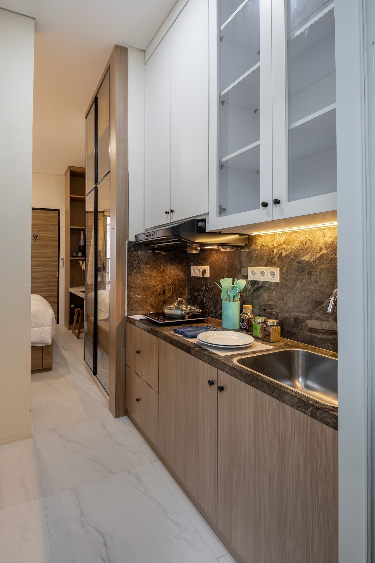 A fully equipped kitchen is displayed with modern cabinetry and a polished marble countertop. Essential appliances, including a stovetop and sink, are positioned for functionality. Nearby, a glimpse of the welcoming sleeping area is visible through a glass partition, enhancing the sense of space.