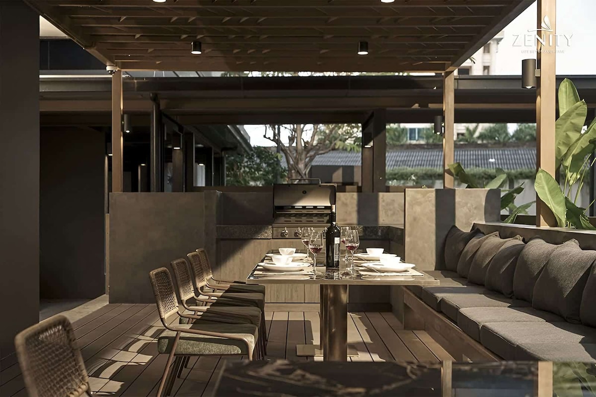 An outdoor dining area features a large wooden table surrounded by cushioned seating and eight chairs. The space is covered with a wooden pergola, providing shade. A grill is visible nearby, and greenery adds a natural touch to the atmosphere.