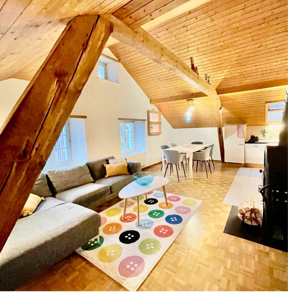 An open living area is highlighted by a sloped wooden ceiling and wooden beams. A comfortable sofa is arranged near a colorful area rug adorned with polka dots. A dining table with chairs occupies one corner, while a kitchen area is visible in the background.