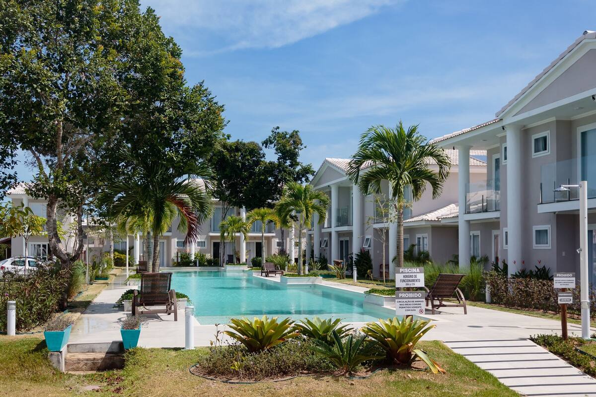 The image showcases a serene resort landscape, featuring a turquoise swimming pool surrounded by palm trees and well-manicured gardens. Modern two-story buildings line the pool area, providing a peaceful atmosphere. Comfortable lounge chairs are positioned around the pool for relaxation.
