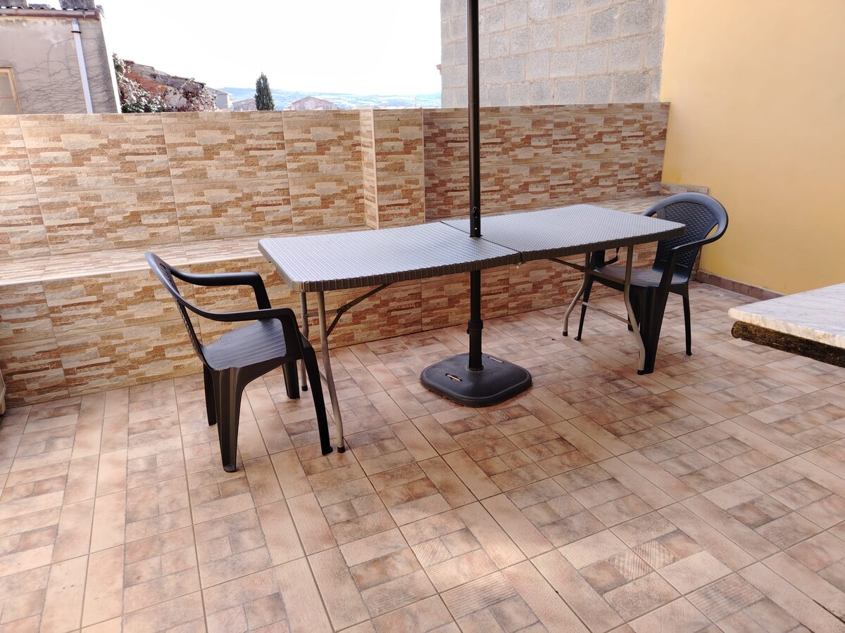 A spacious outdoor area features a rectangular table with an umbrella base. Two black chairs are positioned around the table. Textured stone walls and tiled flooring create a clean and functional space, ideal for relaxation or outdoor dining.