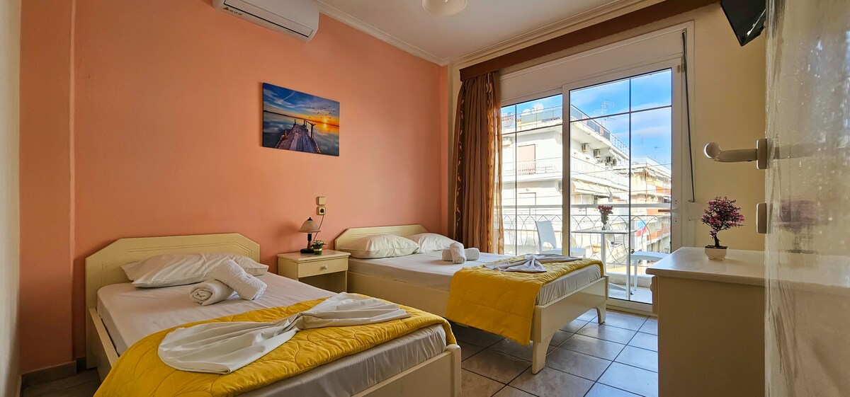 A bright room features two spacious beds, each adorned with clean linens. A small television is mounted on the wall opposite the beds. The window allows natural light to fill the space, while a balcony view of the street and nearby buildings is visible.