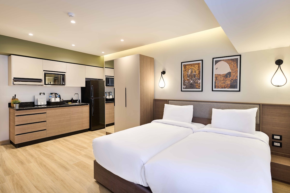 A modern bedroom features two spacious beds with crisp white linens. A stylish kitchenette with dark countertops is visible in the background, complemented by white cabinetry. Light fixtures are mounted on the wall beside the beds, enhancing the contemporary design.