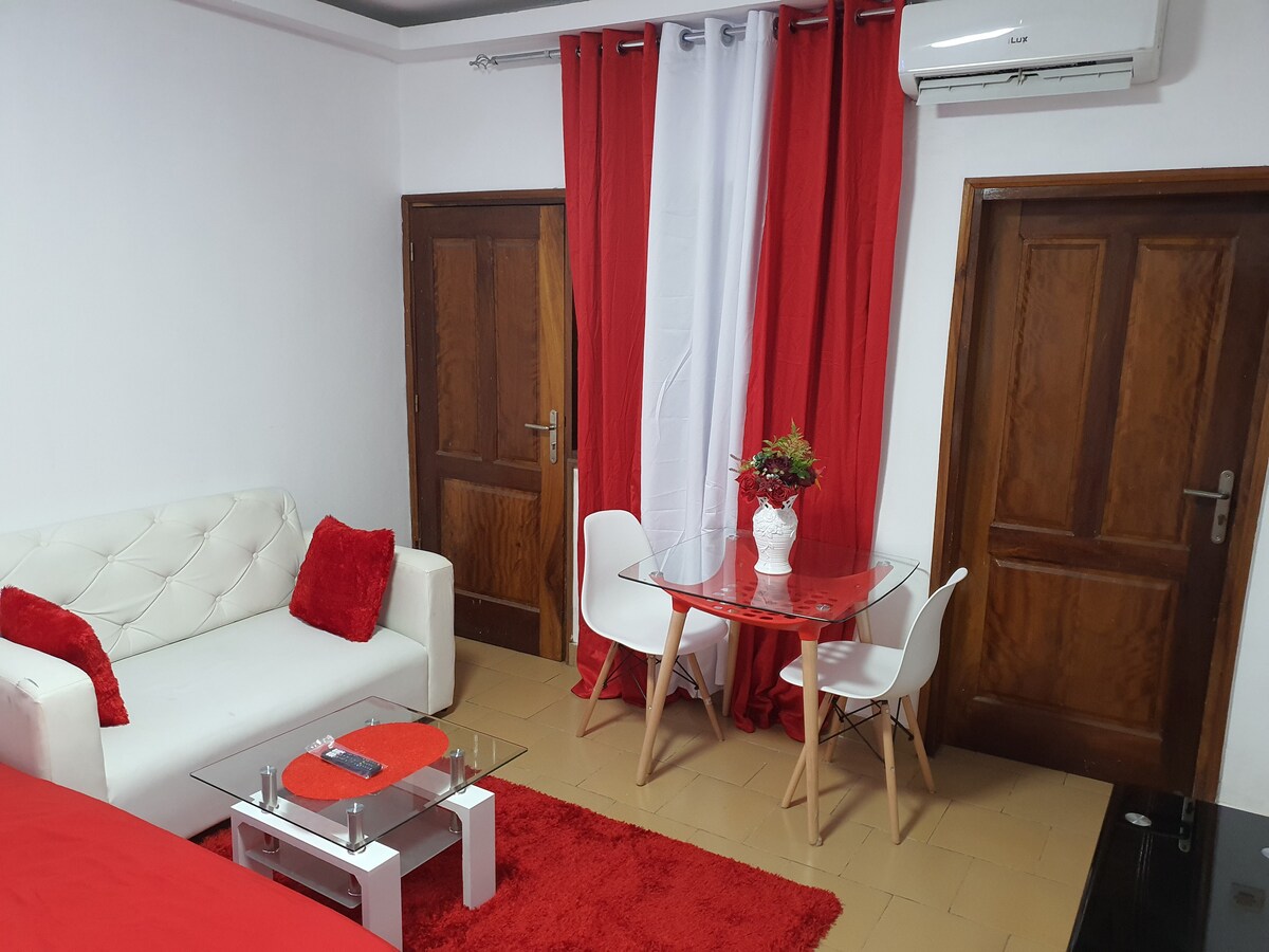 A modern living area includes a white sofa adorned with red cushions, a glass coffee table, and a round dining table with two white chairs. Bright red curtains frame the space, and a small vase of flowers sits on the table, adding a touch of decor.