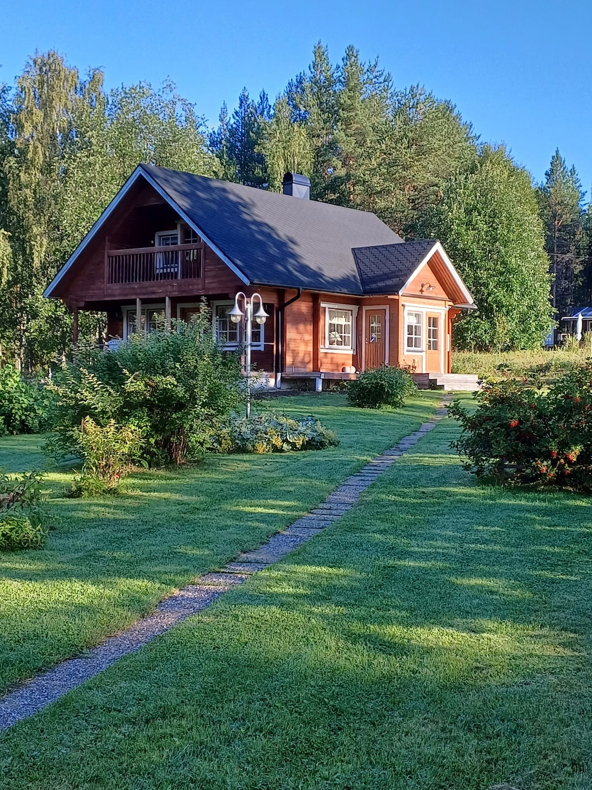 A charming wooden house is framed by lush greenery, with a well-maintained lawn leading to a stone pathway. The exterior showcases a sloped roof and a balcony, inviting a sense of connection with the surrounding nature.