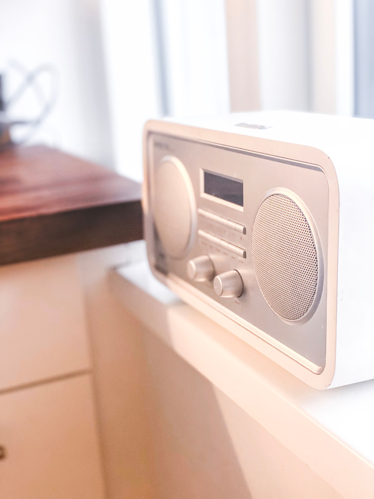 A compact radio is positioned on a windowsill, featuring a sleek silver design. Two dials are visible on the front, along with speaker grilles, providing functionality while enhancing the space.