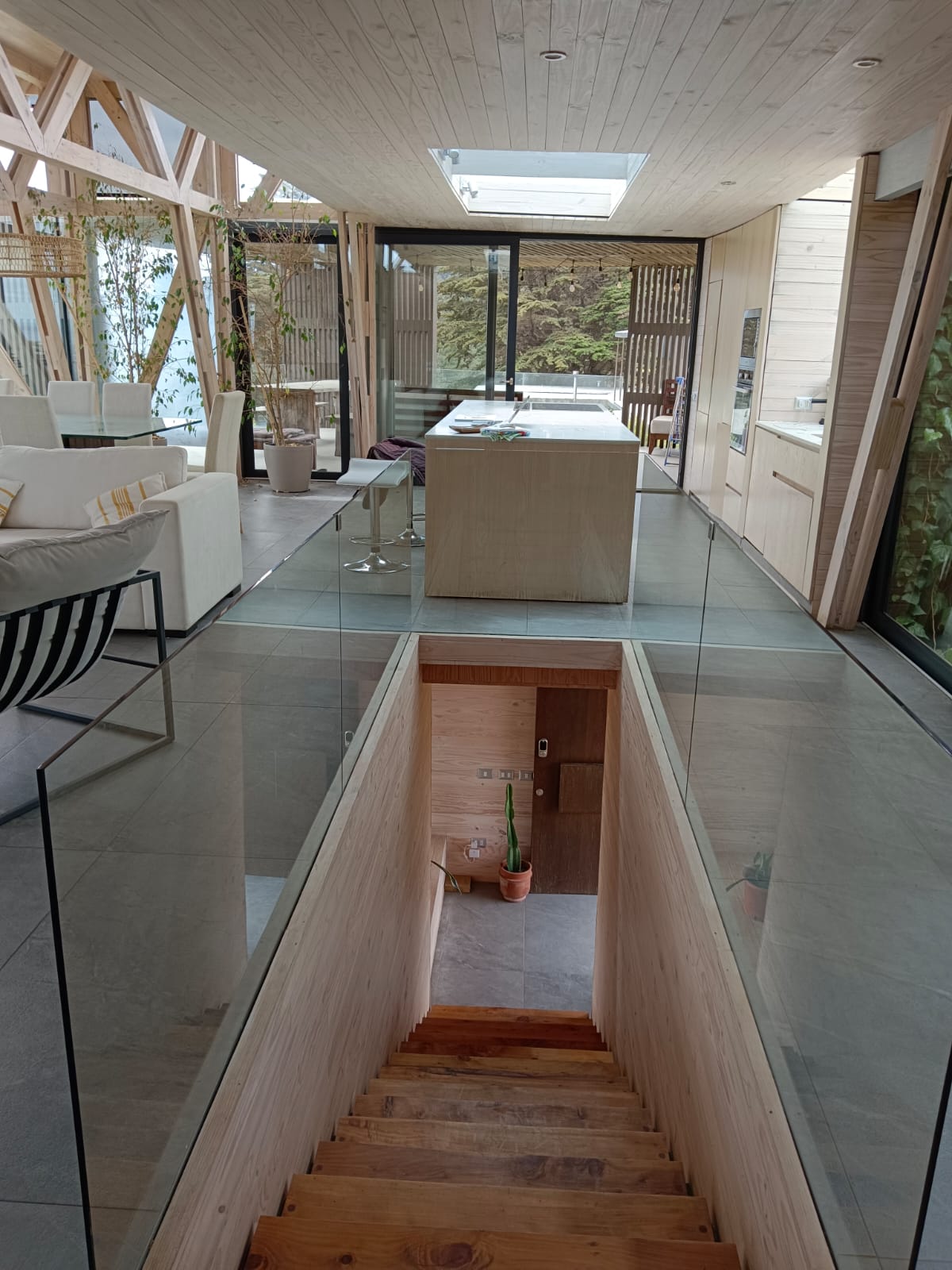 An open and airy space is showcased from an elevated perspective, highlighting a sleek staircase descending to a lower level. Glass railings provide transparency, allowing views of the modern kitchen and living area filled with natural light from large windows.