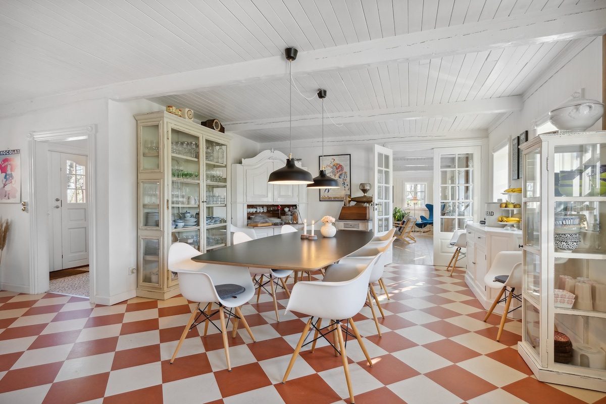 A bright dining area features a large rectangular table surrounded by stylish chairs. The floor showcases a classic checkerboard pattern in red and white. Display cabinets line the walls, holding glassware. Sunlight enters through multiple windows, illuminating the open layout connected to adjacent spaces.