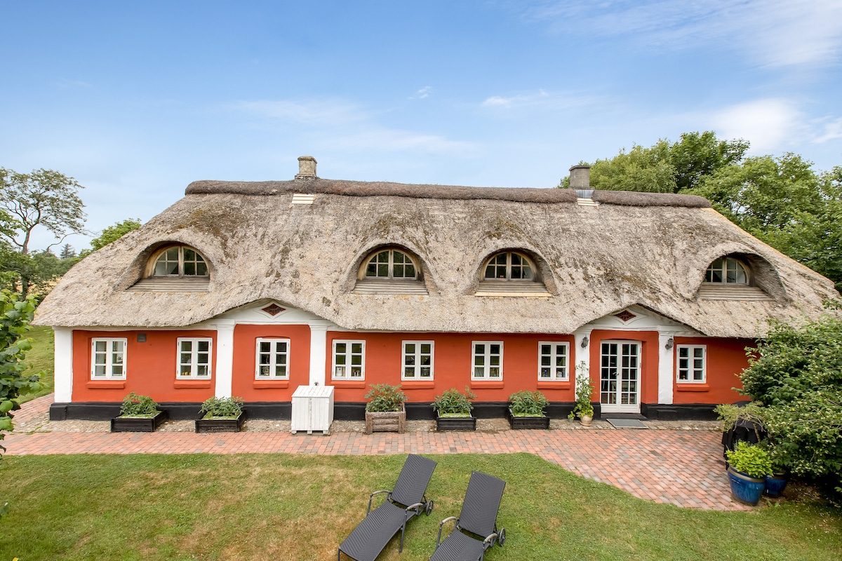 A charming old farmhouse features a classic Danish thatched roof and bright red exterior. Large arched windows are framed with white, enhancing the traditional style. Lush greenery surrounds the building, while patio chairs are positioned on the brick pathway, inviting relaxation in the garden.