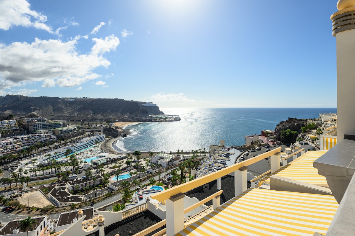 A wide view of the sparkling Atlantic Ocean and the coastline is seen from the terrace. Yellow and white striped awnings provide shade, complementing the landscape. The scene includes palm trees lining the beach and distant mountains under a clear blue sky.