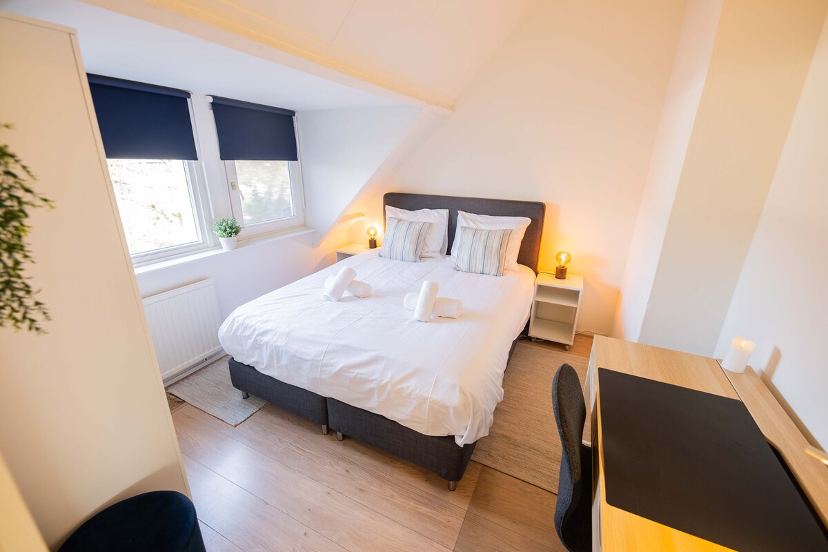 A well-lit bedroom features a spacious bed with crisp white linens and decorative pillows. Two windows with dark roller blinds allow natural light to filter in. A simple desk and chair are positioned against the wall, complementing the minimalist decor.