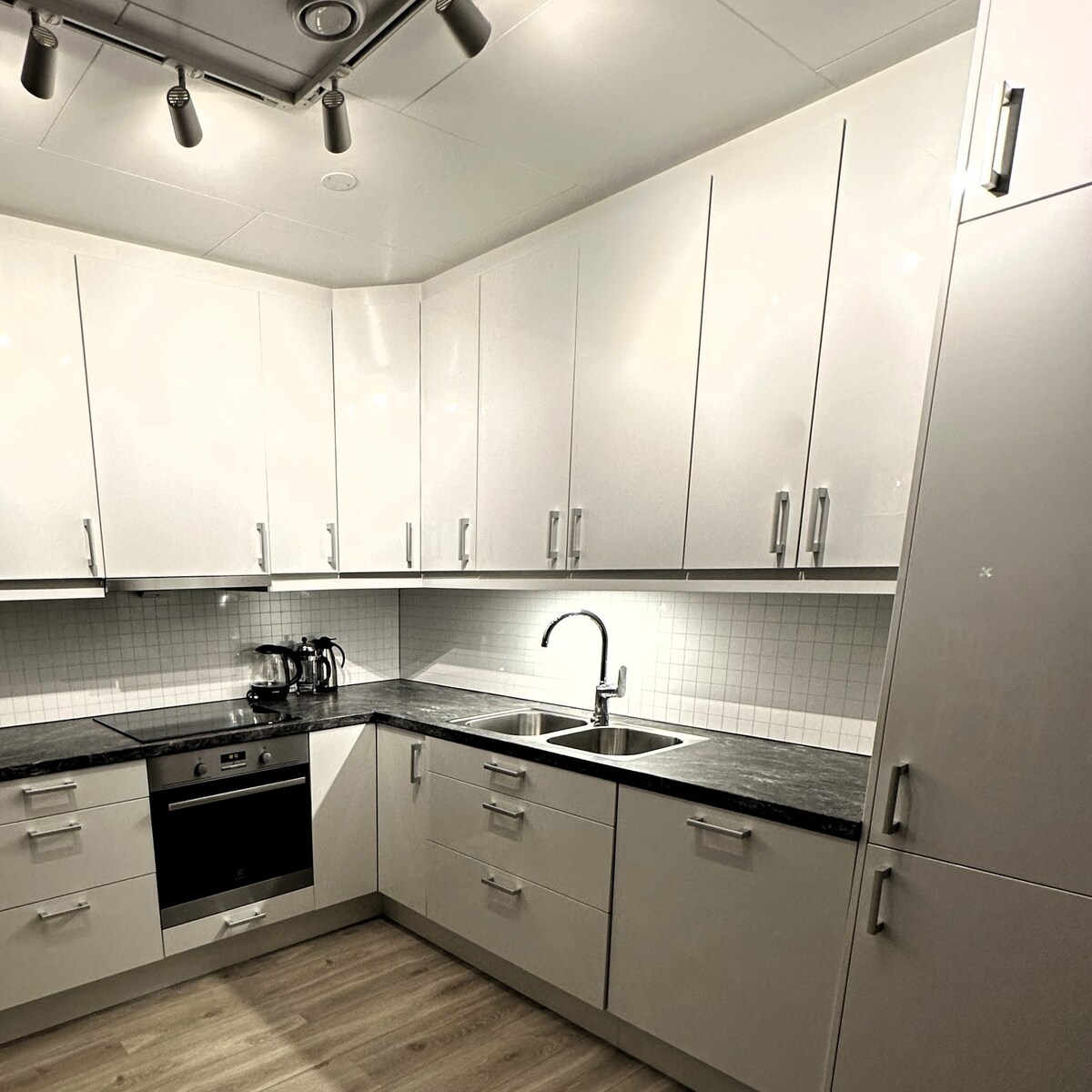 A modern kitchen is presented with sleek white cabinets and a dark countertop. Stainless steel appliances, including an oven and a sink, are integrated into the design. Soft lighting enhances the space, illuminating the organized layout and clean lines of the cabinetry.
