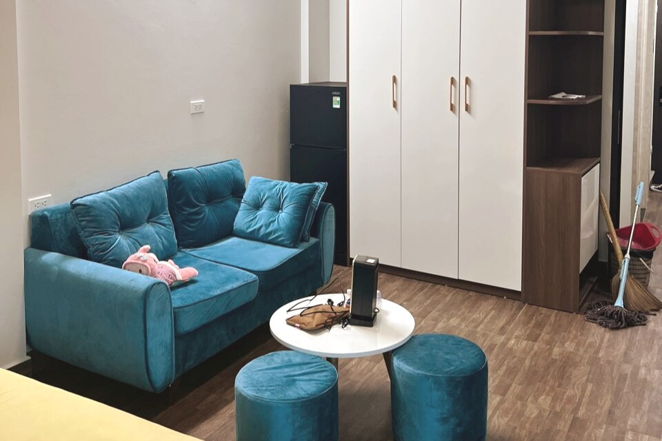 A cozy living area features a blue sofa adorned with plush cushions and a pink stuffed toy. A round coffee table is positioned in front, surrounded by matching ottomans. The space is complemented by a tall wardrobe and a refrigerator in the background.