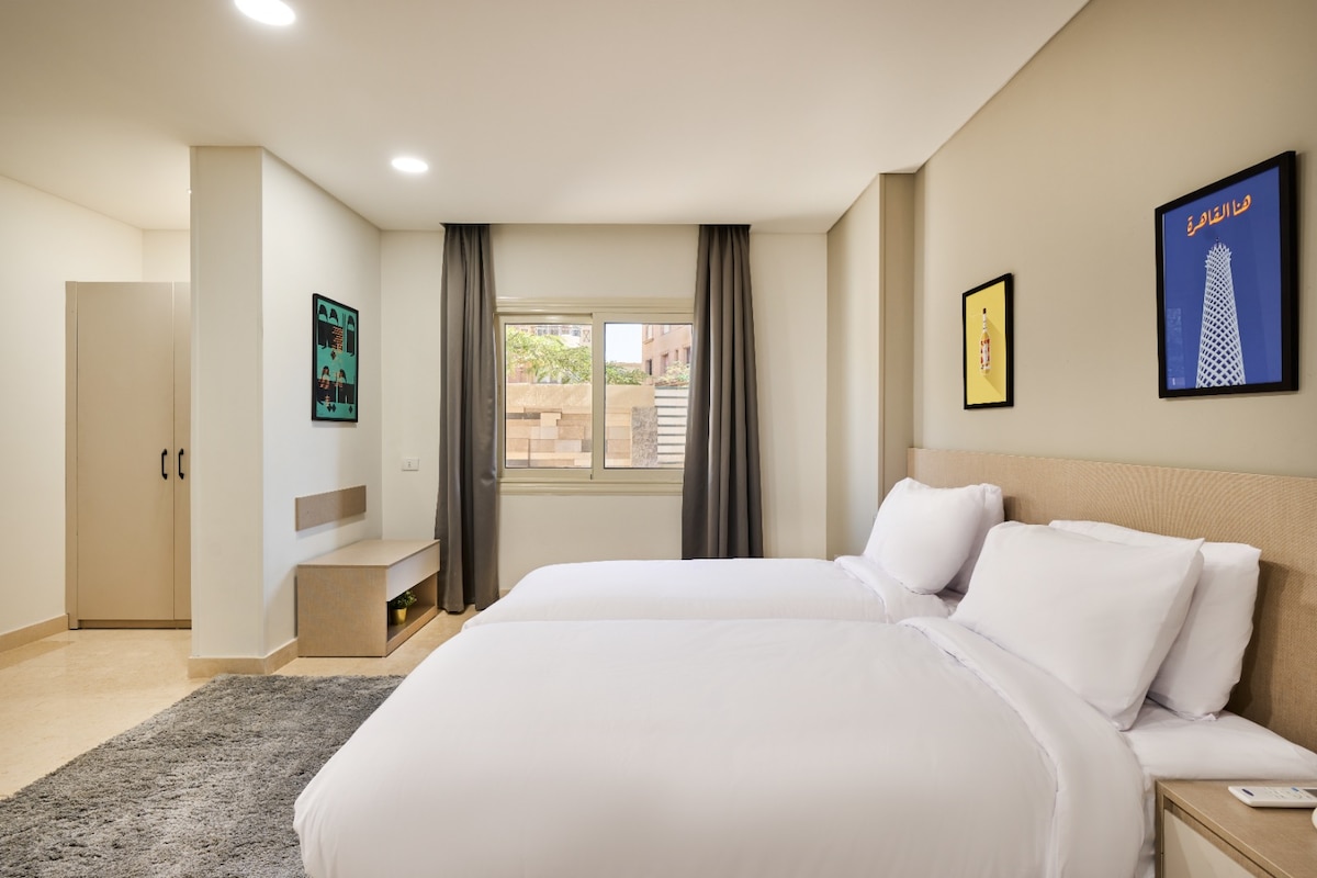 A well-lit guest room features two spacious beds with crisp white linens. A modern wardrobe and a minimalist bedside table offer functional storage. The window overlooks the surroundings, while contemporary artwork adorns the walls, adding a touch of style to the serene space.