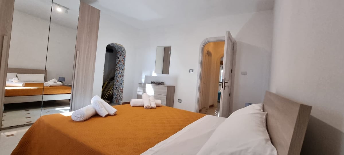 A well-lit bedroom features a spacious bed covered with an orange blanket. White towels are neatly arranged on the bed. A large mirror reflects the room, which includes an arched door leading to another space and a modern dresser against a neutral wall.