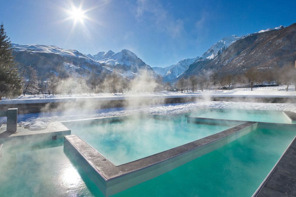 A heated outdoor pool is set against a stunning snowy mountain backdrop. Steam rises gently from the turquoise water, creating a serene atmosphere. The sun shines brightly in the clear sky, illuminating the surrounding landscape.