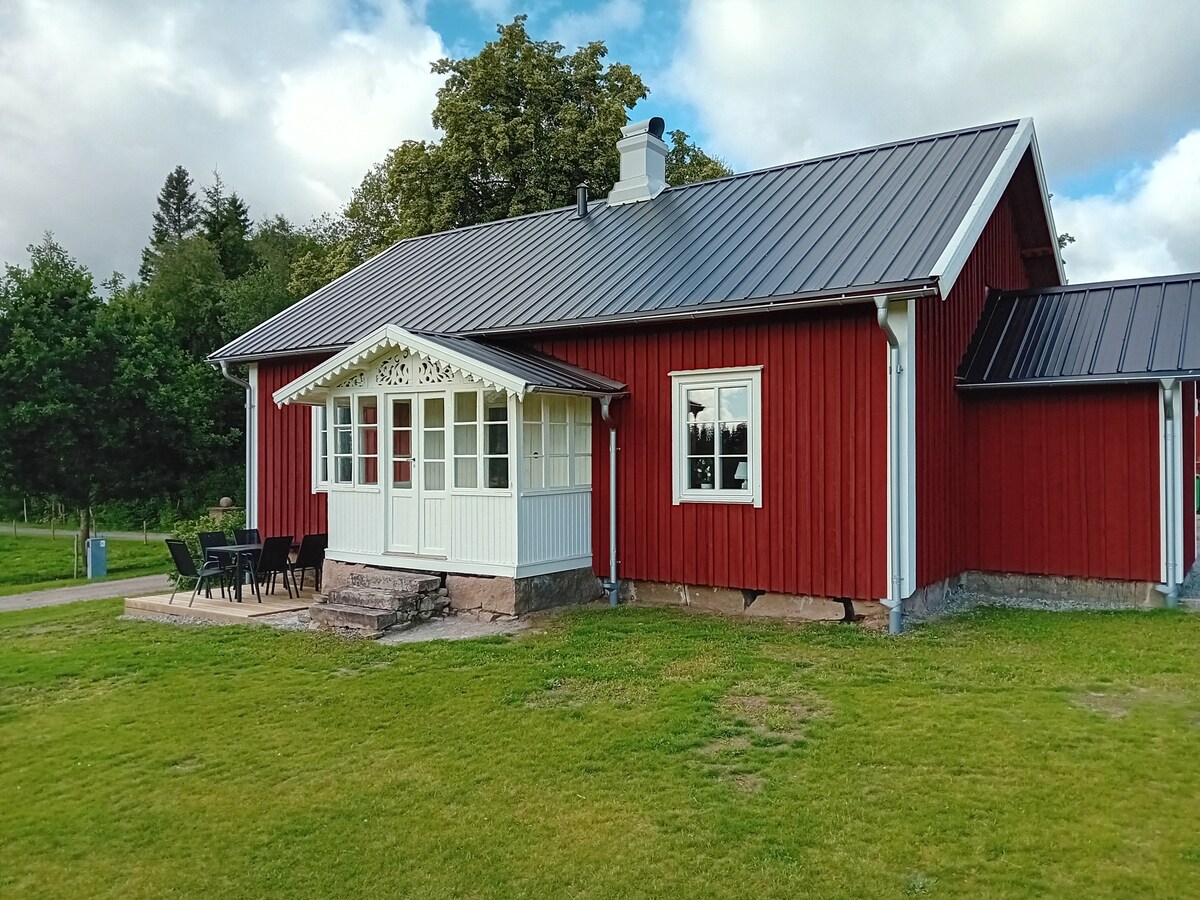 The exterior features a traditional red wooden house with a white accentuated porch. A set of black chairs and a table is positioned on a grassy area, inviting outdoor gatherings. Lush greenery surrounds the property, complementing the calm countryside setting.