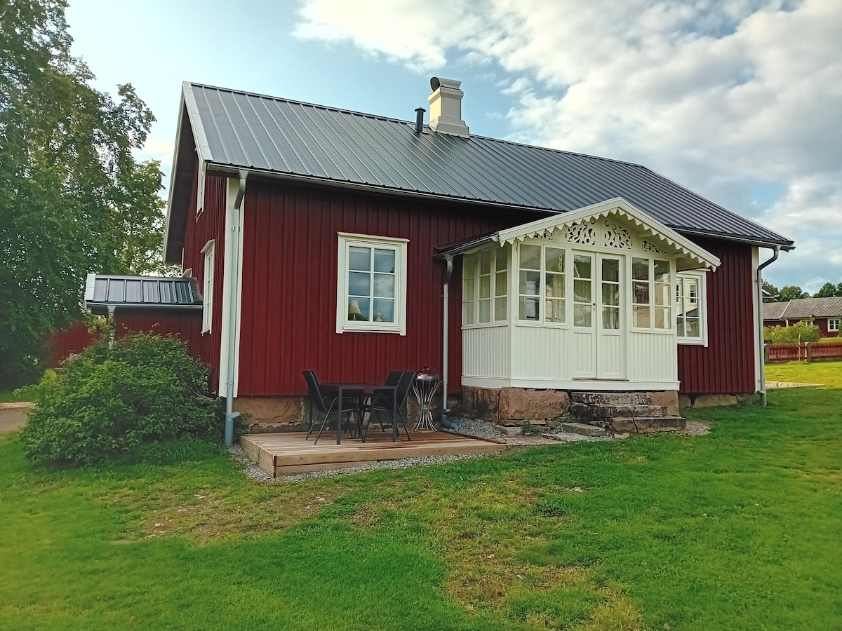 A charming red wooden house features a white sunroom adorned with decorative trim. Adjacent to the house, a small wooden deck with outdoor seating overlooks a grassy area. Large windows provide an inviting connection to the surrounding landscape.