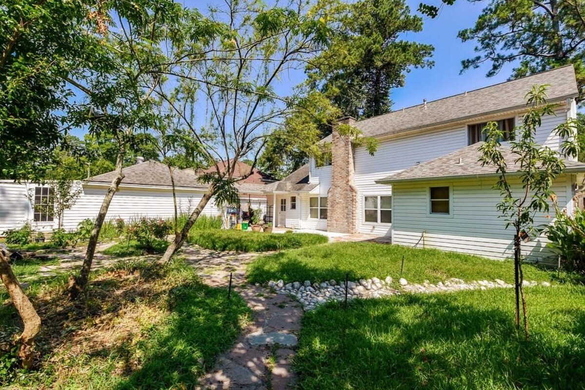 A spacious backyard is surrounded by lush greenery and includes a stone pathway leading to the home's exterior. The house features a stone chimney and multiple windows, allowing natural light to illuminate the outdoor area.