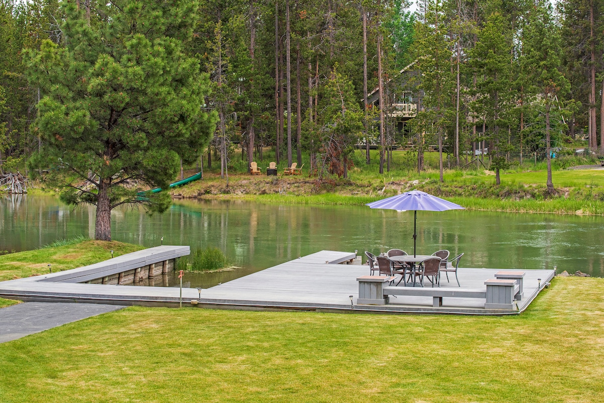 A riverside patio features a shaded dining table surrounded by chairs, positioned on a wooden deck that extends toward the water. Lush green grass surrounds the area, complemented by tall trees in the backdrop, creating a natural and tranquil setting.