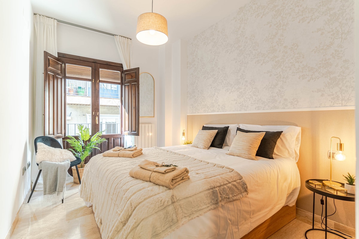 A softly lit bedroom features a spacious bed with neutral bedding and decorative pillows. Two large windows with wooden shutters allow natural light to fill the room. A small side table holds a lamp, while a cozy chair adds an inviting touch to the space.