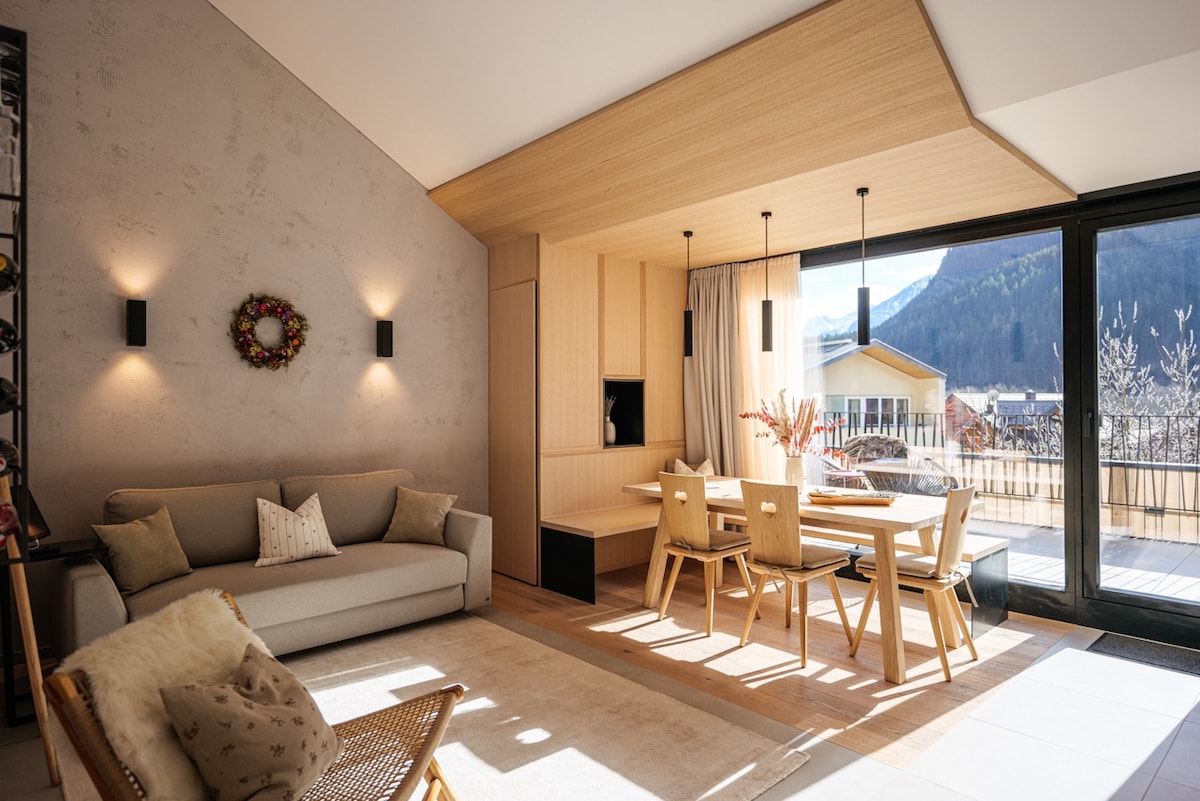 A spacious living area features large windows offering mountain views. A neutral-colored sofa is positioned beside a wooden dining table surrounded by light-toned chairs. Wall-mounted lights provide subtle illumination, and a decorative wreath hangs on the wall, creating a cozy and inviting environment.