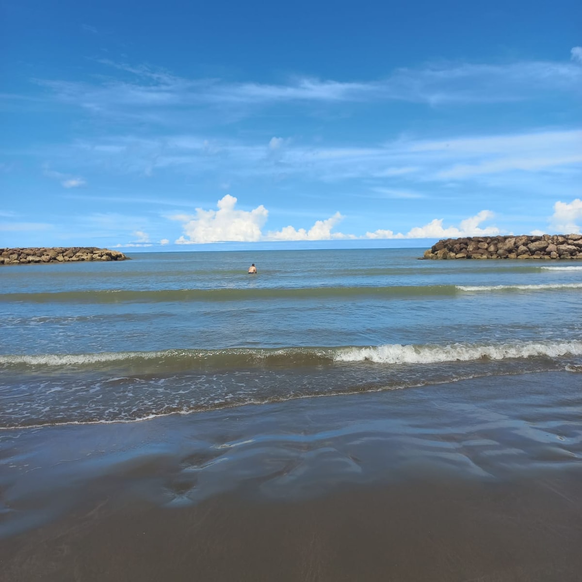 The image captures a serene beach scene featuring gentle waves lapping at the shore. Two rock formations are visible in the distance, framing the horizon where the sky meets the sea. A lone person is seen wading in the water, contributing to the tranquil atmosphere.