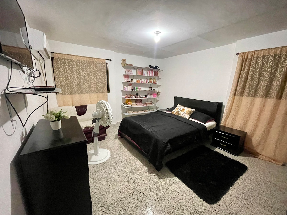 A cozy room features a spacious bed with dark bedding and a decorative pillow. Shelving displays various personal items. A fan provides cooling, while a television is mounted on the wall. Light curtains frame the window, and a plush rug adds warmth to the concrete floor.
