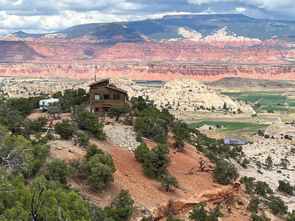 A cabin is nestled among rocky outcrops and juniper trees, situated on a plateau. Vast desert landscapes extend behind it, showcasing layered red and orange rock formations under a partly cloudy sky. The serene environment reflects a sense of isolation and natural beauty.