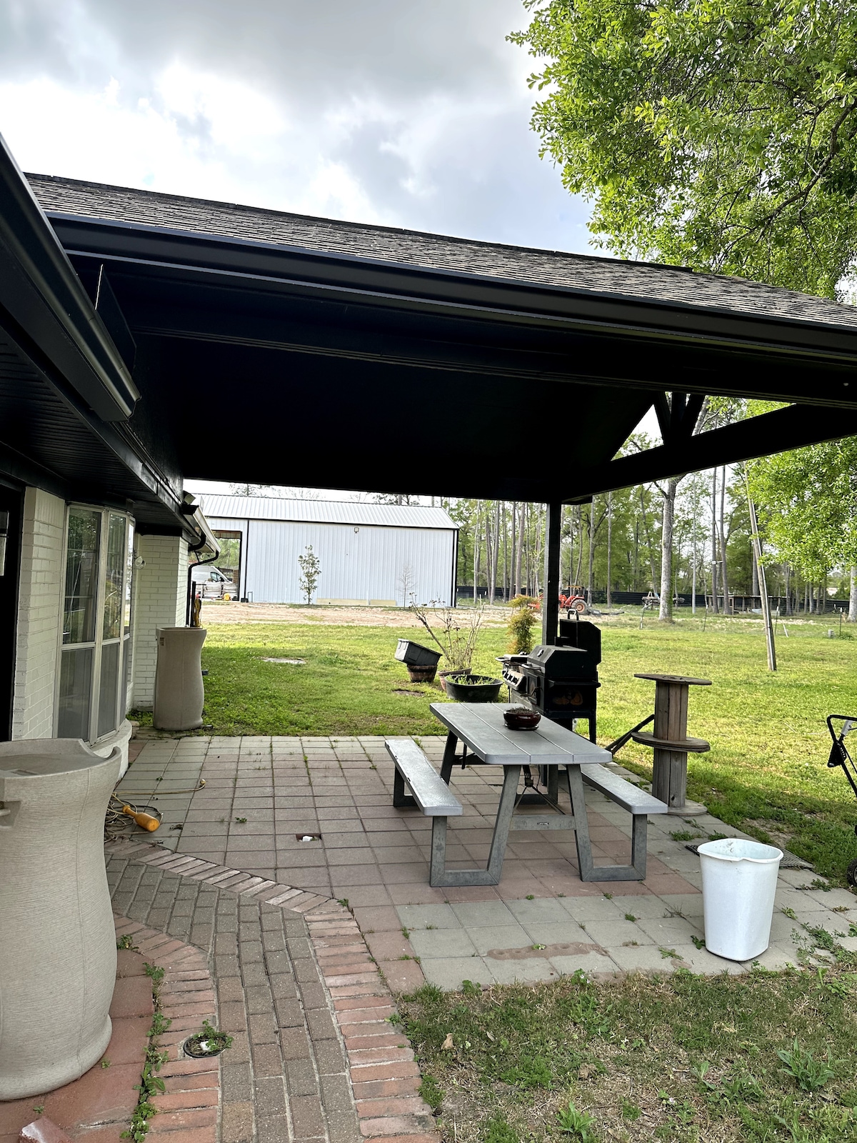 A covered outdoor patio features a rectangular picnic table surrounded by several benches. A grill is positioned nearby, and the space includes a paved pathway and grassy areas. A large building is visible in the background, along with trees providing a natural setting.