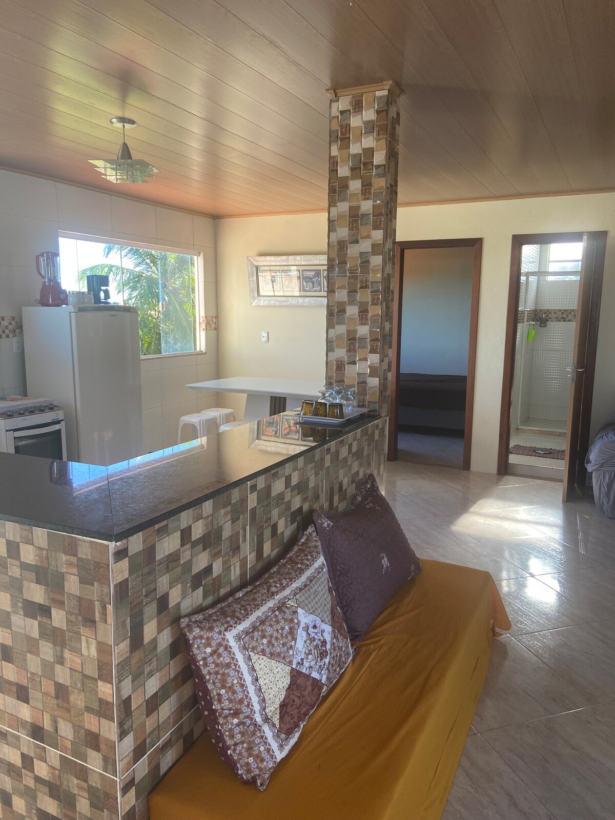 A modern kitchen area is visible, featuring a granite countertop with a mosaic tile backsplash. Nearby, a seating space with patterned cushions offers a comfortable spot. Large windows allow natural light to fill the room, with a glimpse of greenery outside.