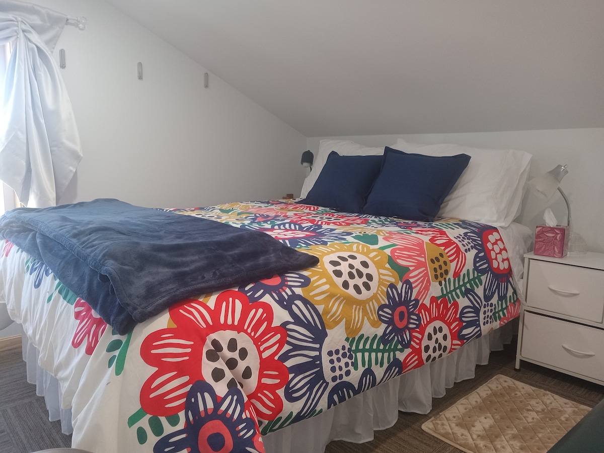 A bright sleeping area features an extra long full bed adorned with a colorful floral comforter and plush navy pillows. Soft lighting is provided by a bedside lamp, and a small nightstand holds personal items. Natural light streams through a nearby window with sheer curtains.