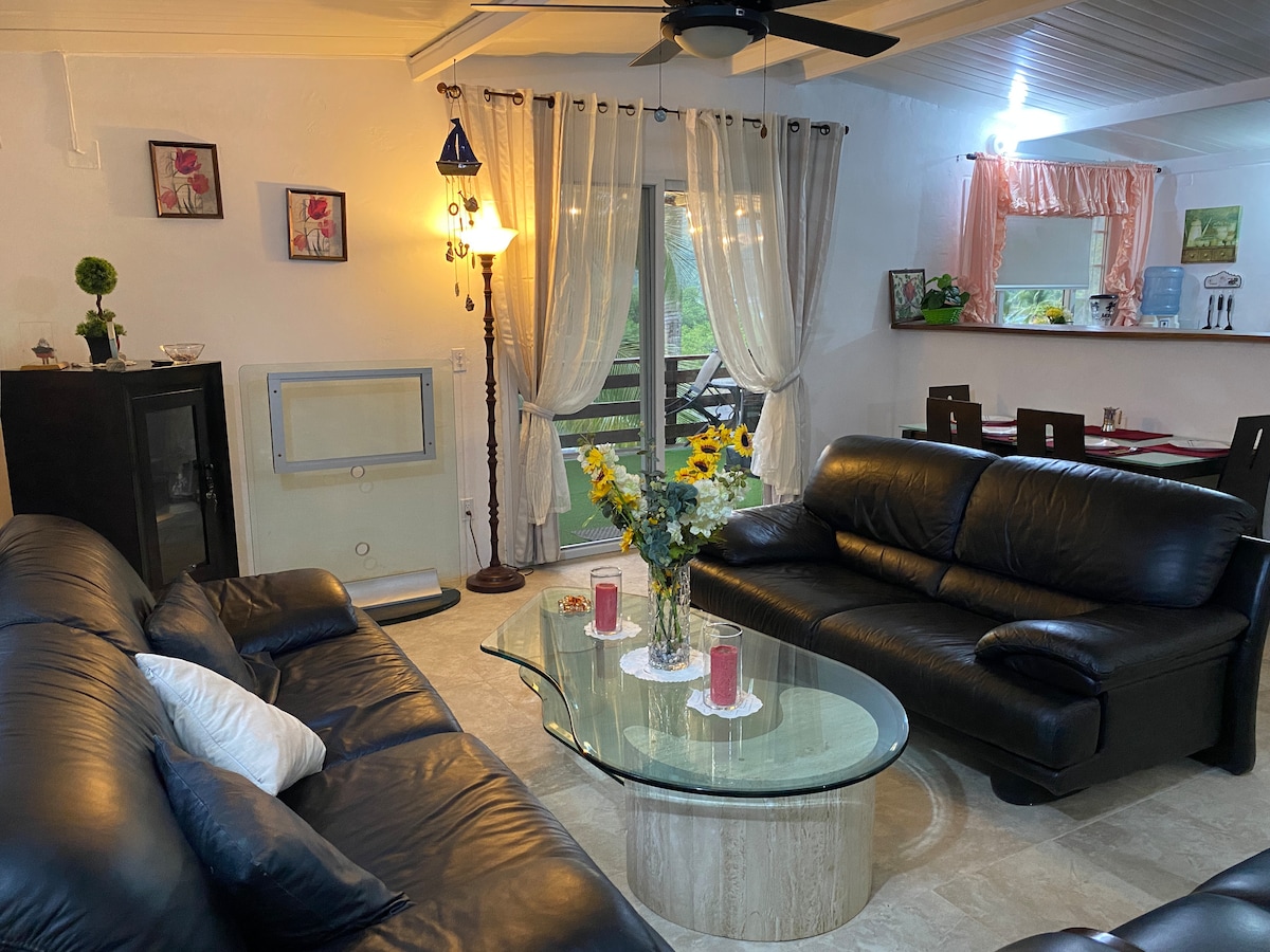 An air-conditioned living room features two black leather sofas facing a glass coffee table, which holds arrangement of flowers and candles. Natural light enters through the large windows adorned with sheer curtains, while a dining area is visible in the background.