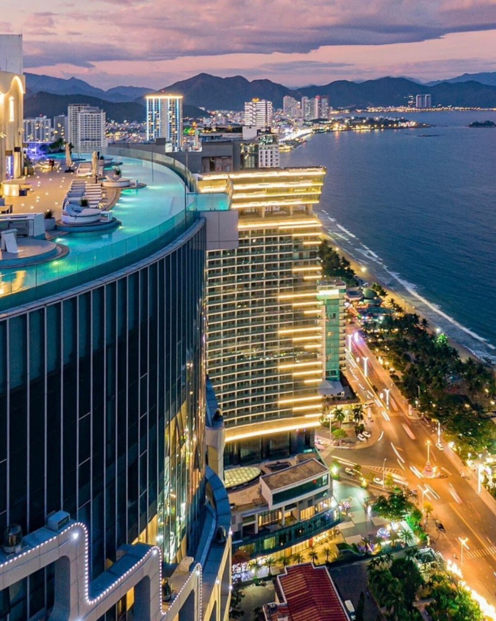 An aerial view captures the coastline and city skyline of Nha Trang at dusk. The illuminated buildings reflect a vibrant lifestyle, while the ocean glistens under the soft light of the setting sun. Rooftop amenities are visible, offering insights into the location's lively atmosphere.