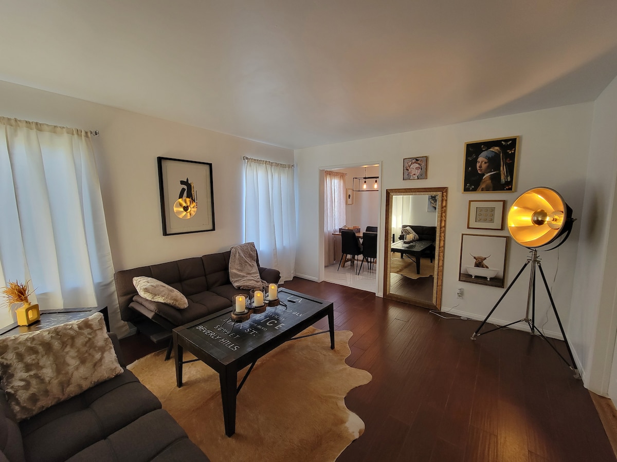 A modern living area is highlighted by a comfortable sofa and a sleek coffee table, surrounded by natural light from large windows. Stylish decor, including a floor lamp and wall art, complements the warm tones of the wood flooring.