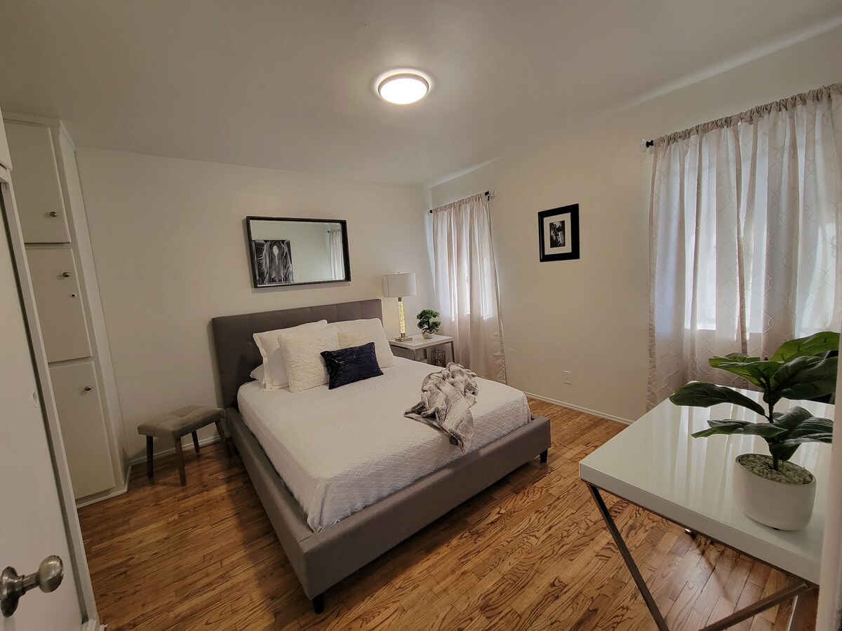 The bedroom features a spacious bed dressed in white linens, accented with a dark cushion. A small chair is positioned nearby, and a modern desk with a plant is present. Light streams through sheer curtains, illuminating the warm wooden floor.