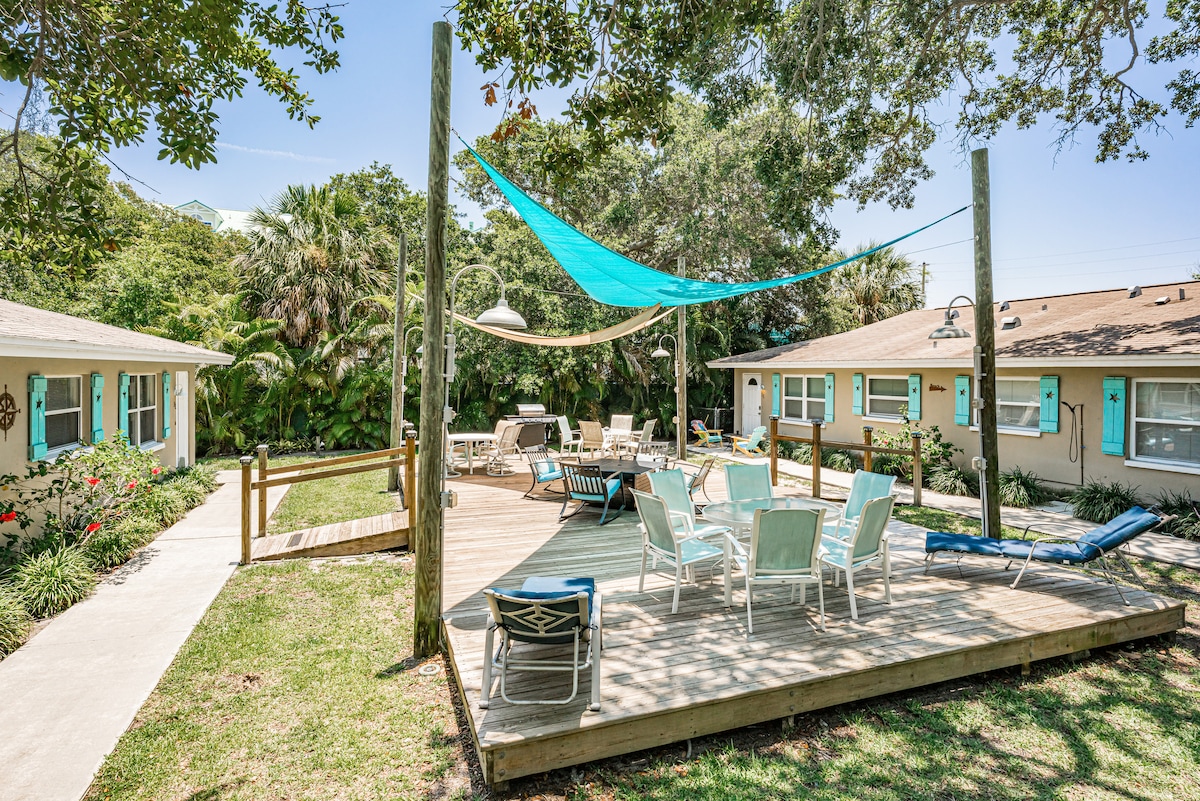 A spacious outdoor deck is surrounded by greenery, featuring multiple seating areas. A large blue shade sail provides coverage over chairs and loungers. The deck flows into a landscaped area with flowering plants, creating a serene environment for relaxation and socializing.