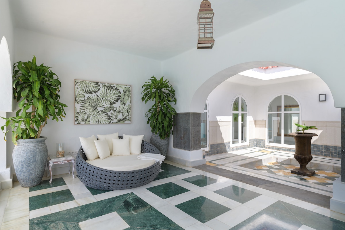 A spacious outdoor area features a round, wicker lounge bed adorned with soft cushions. Decorative plants are positioned throughout, while a stone column fountain adds a tranquil touch. Sunlight floods the space through arched openings, creating a bright and inviting atmosphere.