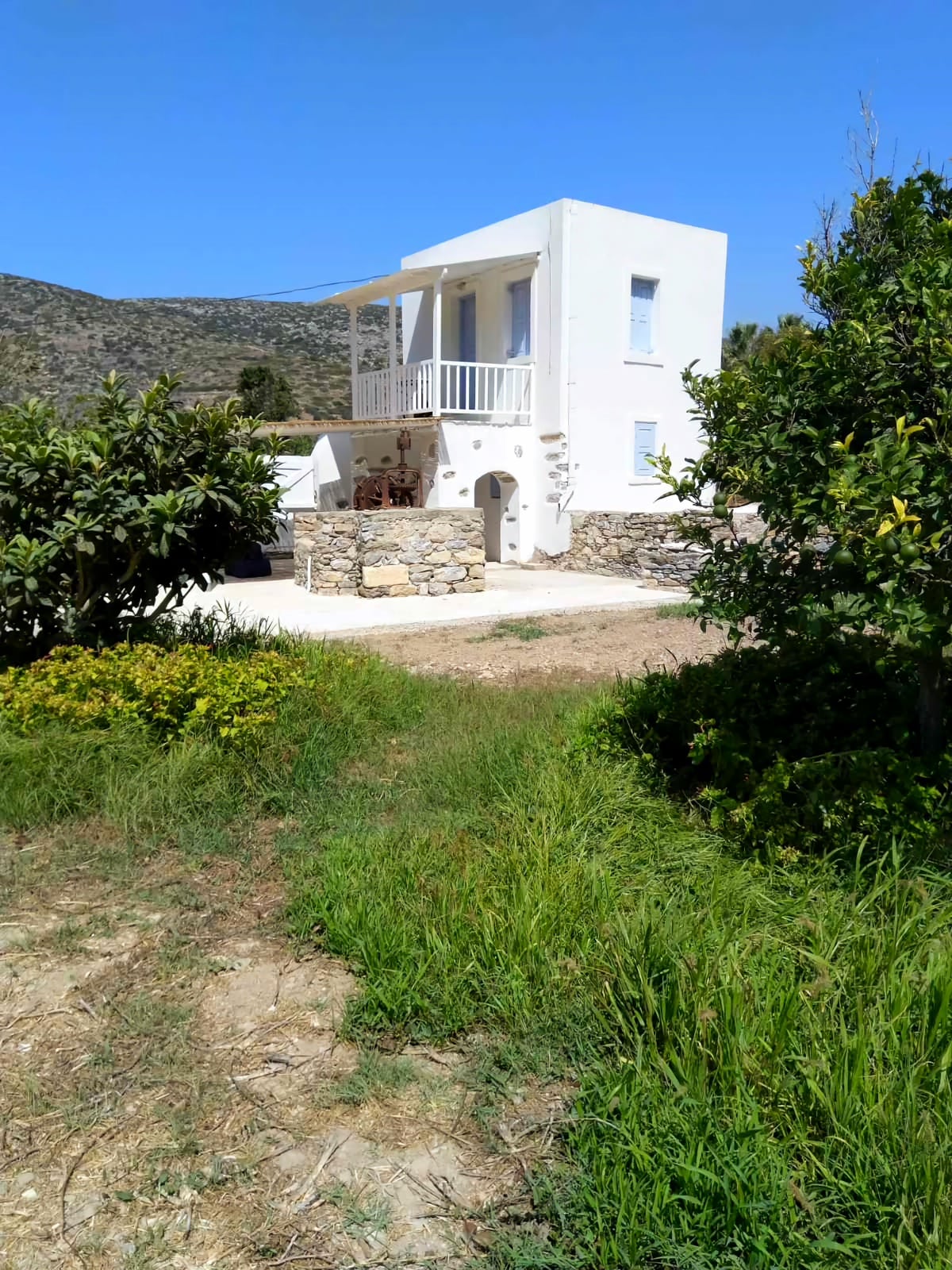 The two-story country house is presented surrounded by a lush green landscape. The exterior features white walls and a stone foundation, complemented by a small balcony on the upper level. Nearby, foliage and plants contribute to the serene ambiance of the estate.