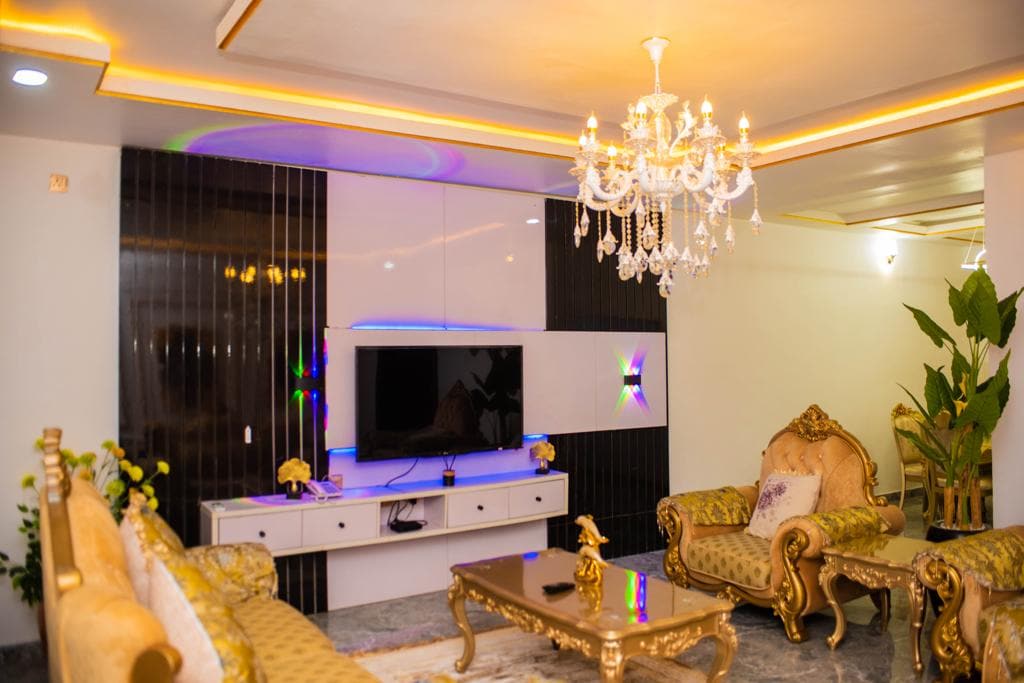 A spacious living area is highlighted by an elegant chandelier and a stylish seating arrangement with golden accents. A modern television is mounted on the wall, complemented by a decorative console. Indoor plants add a touch of freshness to the environment.
