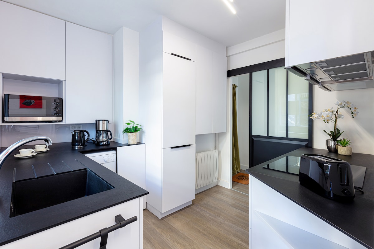 A modern kitchen is presented with sleek white cabinetry and dark countertops. Appliances include a coffee maker, kettle, and toaster. Natural light enters through a glass door, while a potted plant adds a touch of greenery to the space.