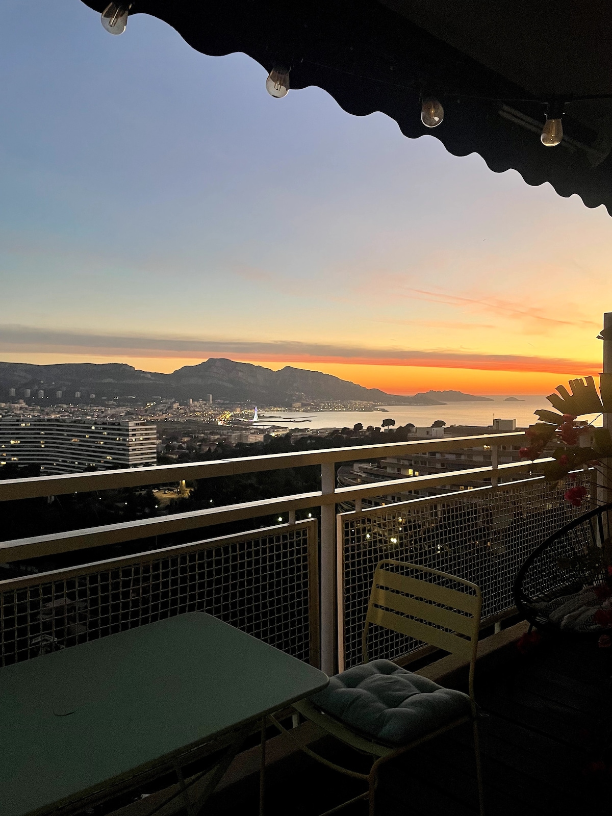 A balcony provides a panoramic view of the sea and surrounding landscape during sunset. The sky is filled with warm hues, and the distant mountains frame the horizon. A small table and chairs are positioned on the balcony, inviting outdoor relaxation.