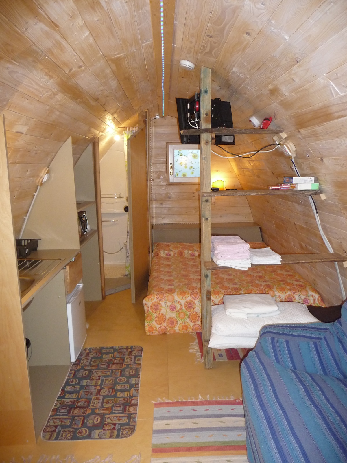 An interior view of a wooden bungalow showcases a cozy sleeping area with a mattress covered in a patterned blanket. A small kitchenette with basic appliances is visible, alongside a space for relaxation furnished with a striped sofa. Light illuminates the warm wooden walls.