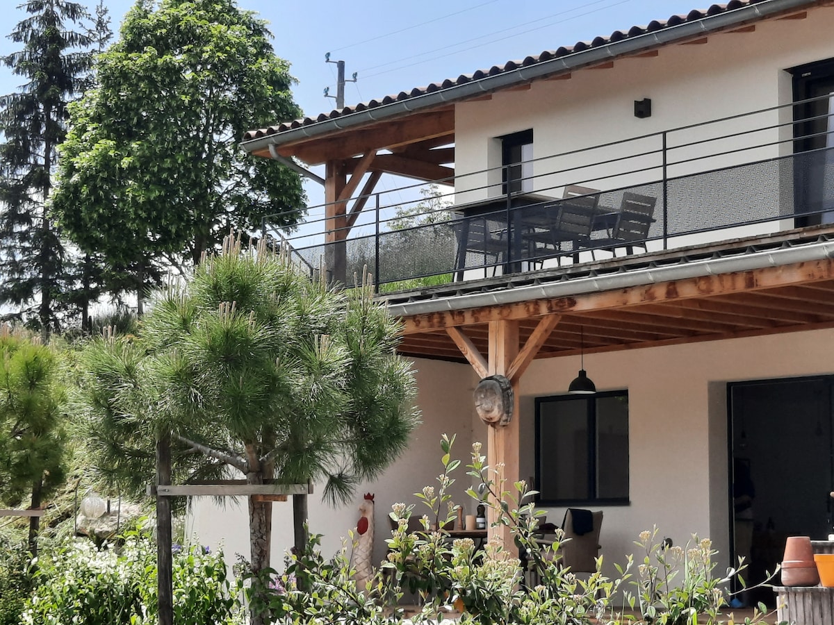 The image shows a wooden terrace with outdoor furniture, including a dining table and chairs, positioned under a roof extension. Surrounding greenery features neatly trimmed shrubs and a potted plant. The structure integrates natural elements with a modern design, highlighting the outdoor living space.