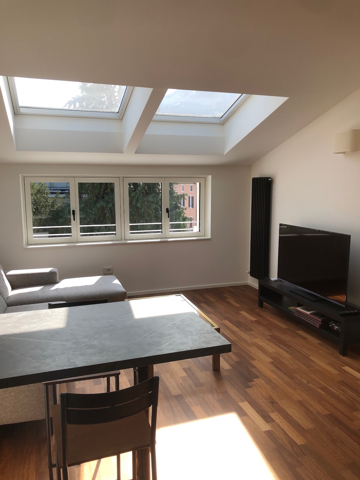 A bright living area features a cozy sofa situated near large skylights that allow natural light to flood the space. A dining table with chairs is positioned in front, while a flat-screen television is mounted on the wall, adding functionality to the room.