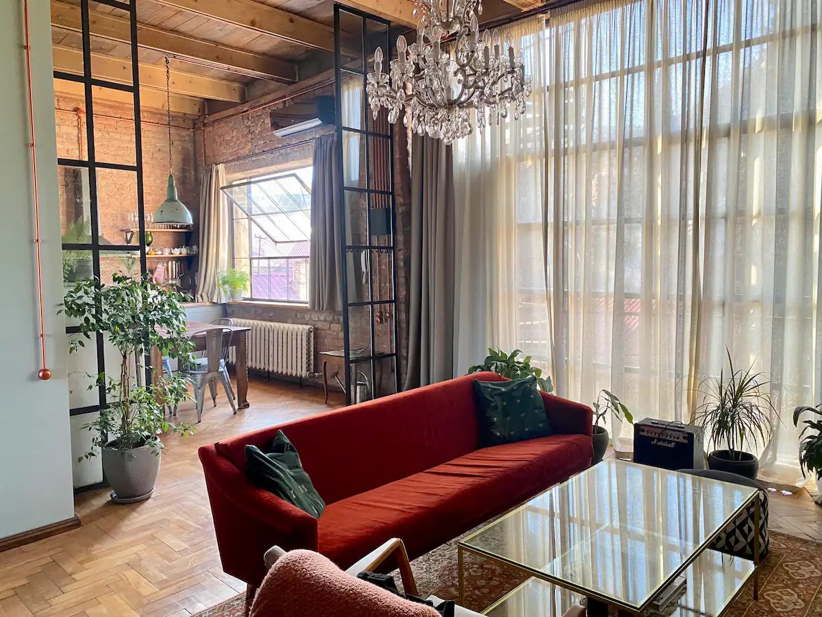 The living area showcases a spacious red sofa complemented by decorative pillows. A vintage chandelier hangs above a glass coffee table. Large windows allow natural light to fill the room, creating a bright atmosphere. Potted plants add green accents, enhancing the rustic decor.
