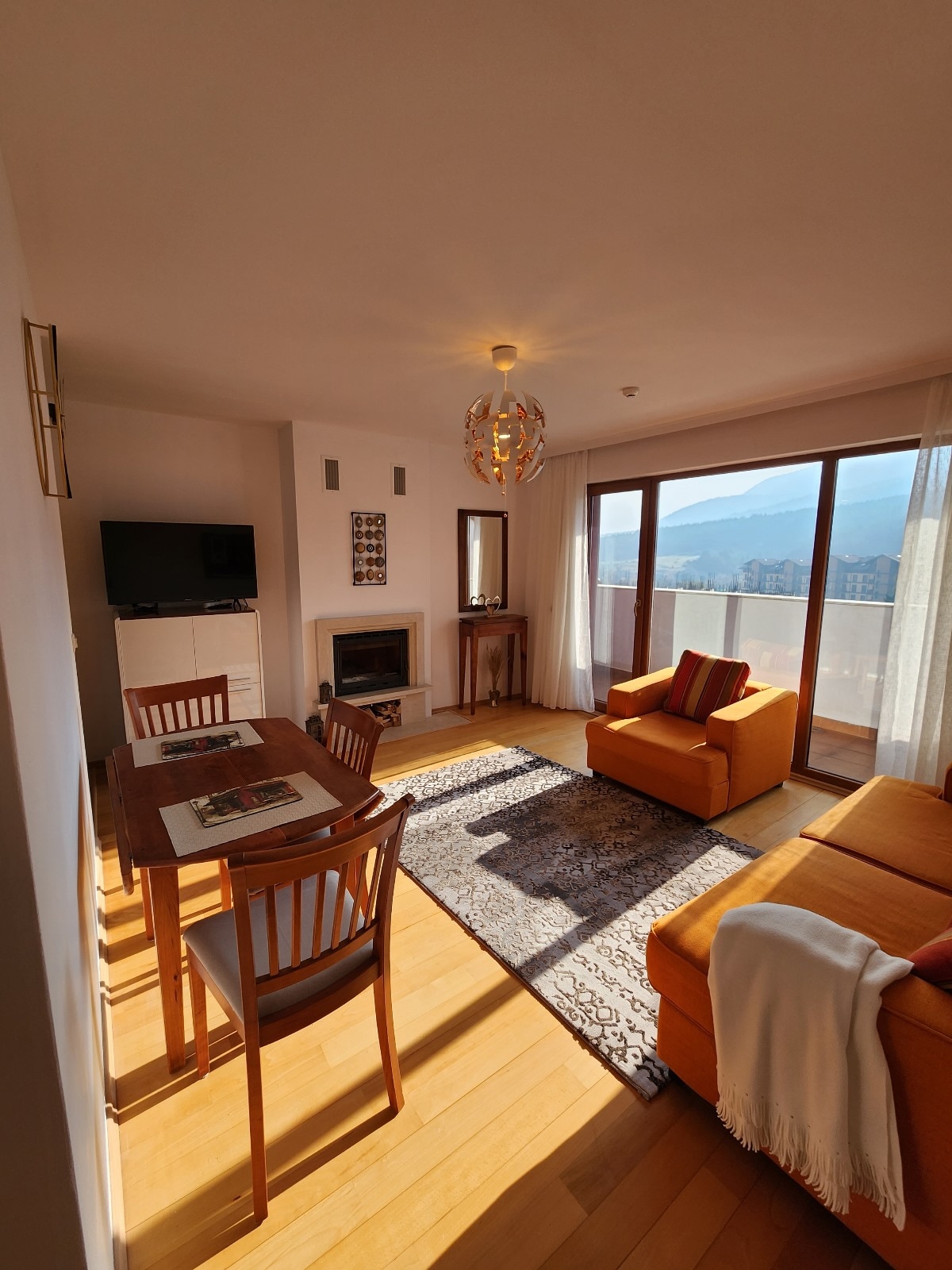 A sunlit living area features hardwood floors and large glass doors leading to a balcony with mountain views. The space includes a cozy orange sofa, a dining table for four, and a flat-screen TV, complemented by a fireplace and decorative accents.