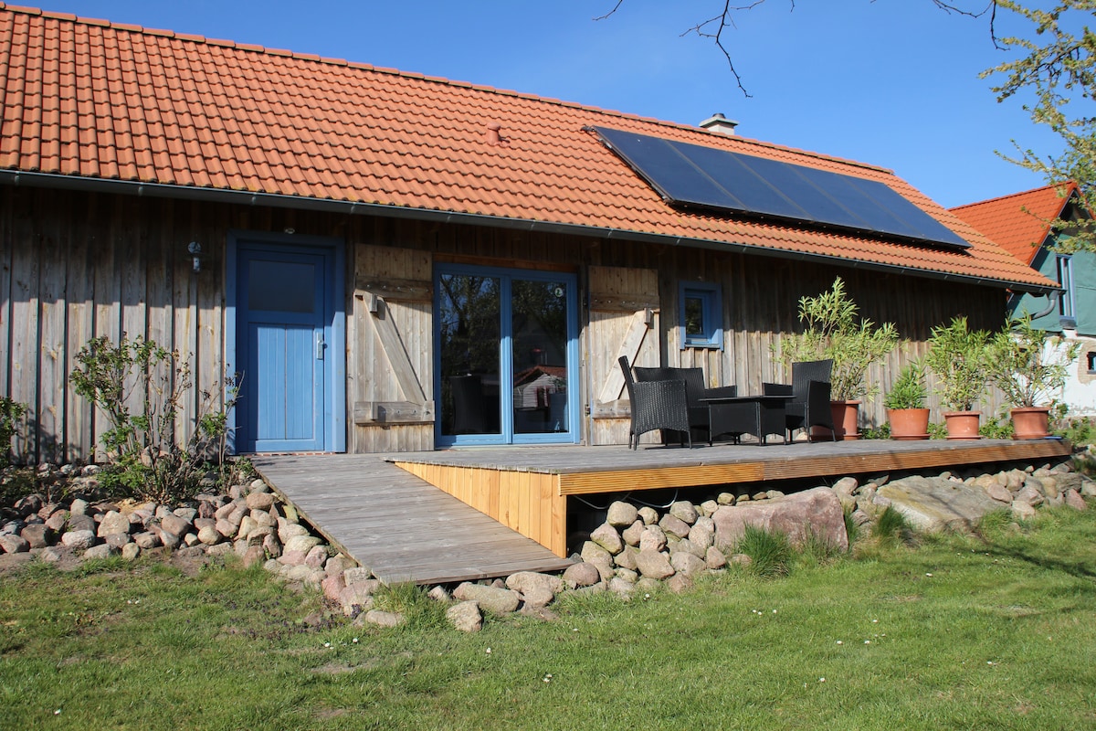 The exterior of a wooden bungalow is presented, showcasing a sloped roof with solar panels. A deck area features outdoor seating and potted plants, surrounded by a rocky landscape. The entrance includes a bright blue door, enhancing the rustic charm of the structure.