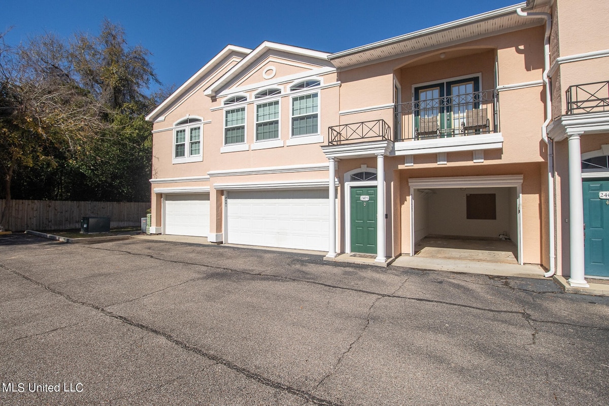 The exterior of a two-story condominium building is displayed, showcasing a light peach facade. Two garage doors are visible on the ground floor, flanked by green double doors leading to a balcony on the second level. A paved driveway extends in front.